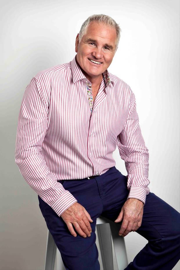 Man in pink striped shirt and navy pants, smiling, seated on a stool, studio background.