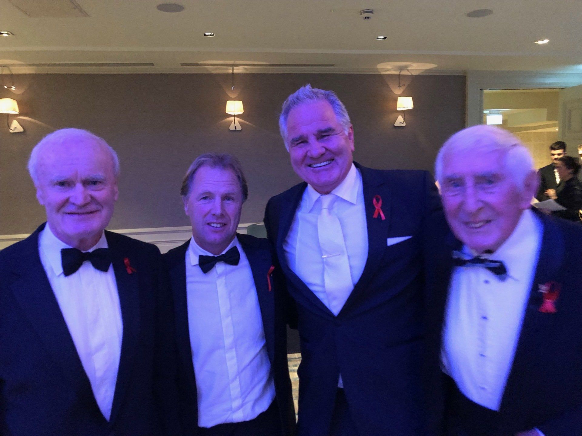 Four men in tuxedos with red ribbons, smiling in a room with sconces.