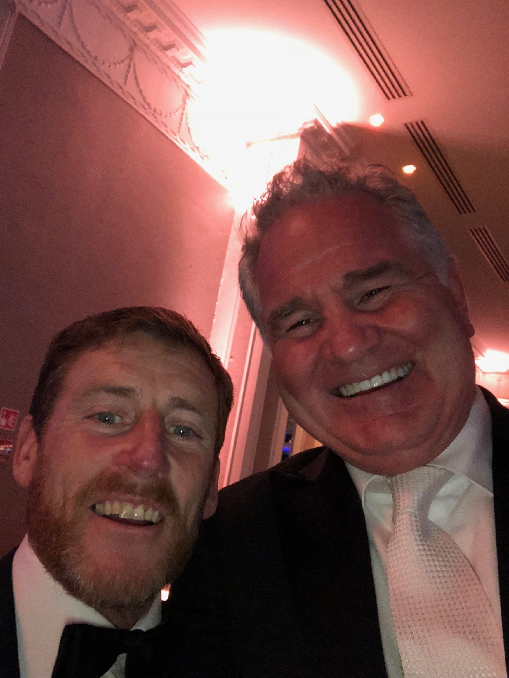 Two men smiling at a camera, indoors. One has a beard, the other wears a tie. Bright ceiling light.