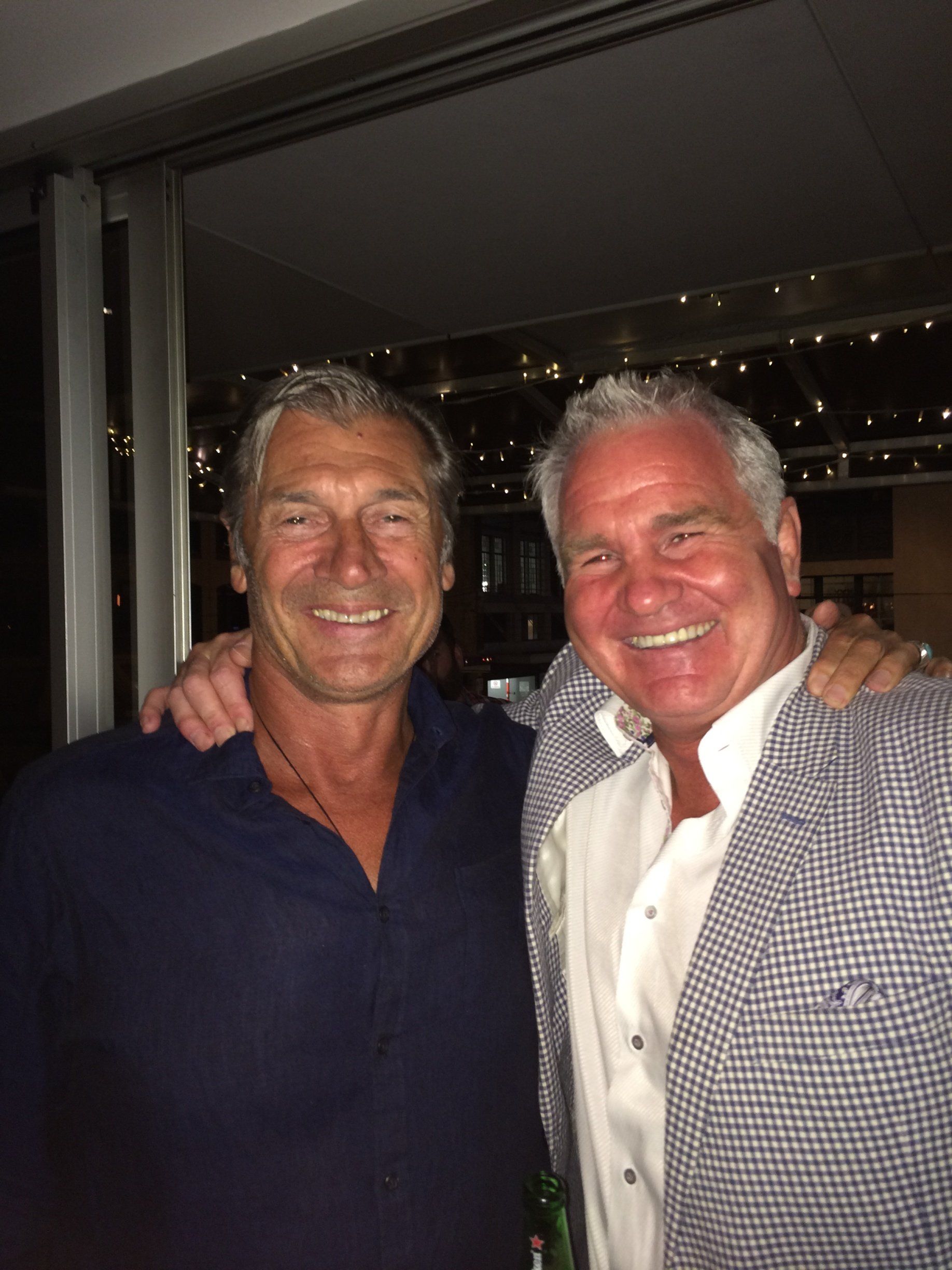 Two men smiling, arm in arm. One in a blue shirt, the other in a white shirt and patterned blazer. Lit background.