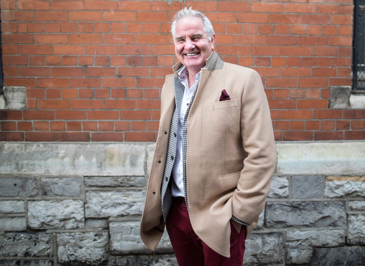 Man in a tan jacket, paisley shirt, and burgundy pants smiles in front of a brick and stone wall.
