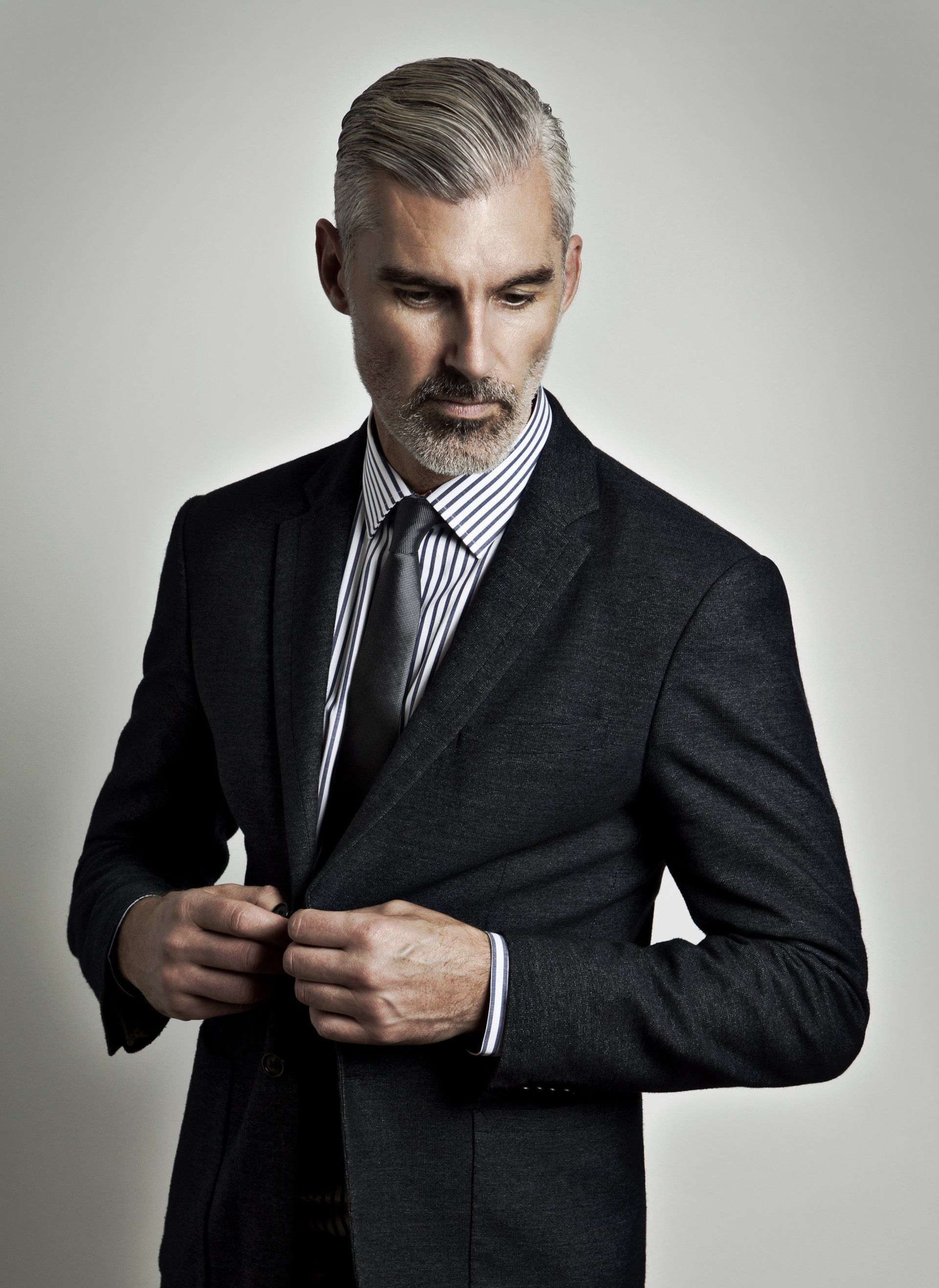 Man in a dark suit, buttoning his jacket, looking down. Gray hair, striped shirt, neutral background.