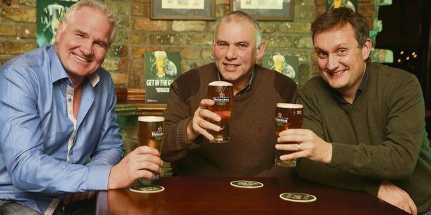 Three men smiling, toasting with pints of beer in a pub setting.