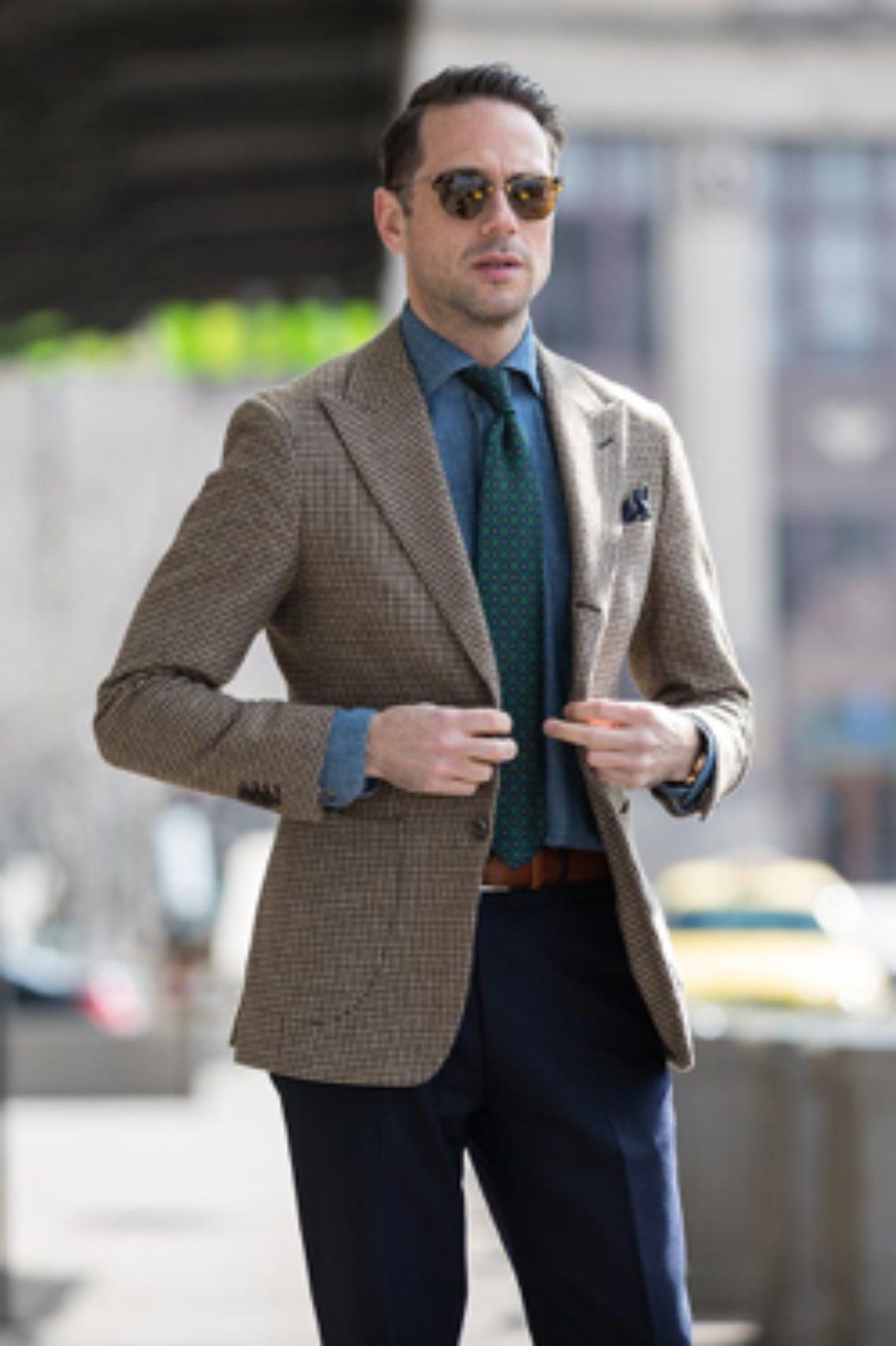 Man in a tweed blazer, blue shirt, tie, and navy pants, outdoors, sunny day.