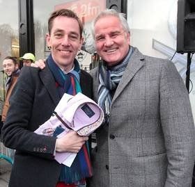 Two men smiling, one holding a shirt; outdoors. They wear coats and scarves.