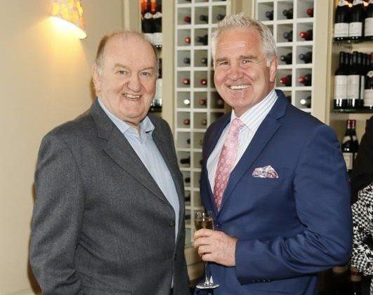 Two men smiling, one in a blue blazer with a pink tie and the other in a grey suit, standing near a wine rack.