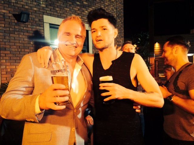 Two men arm-in-arm outside at night, holding beer. One in a suit, the other in a black tank top.