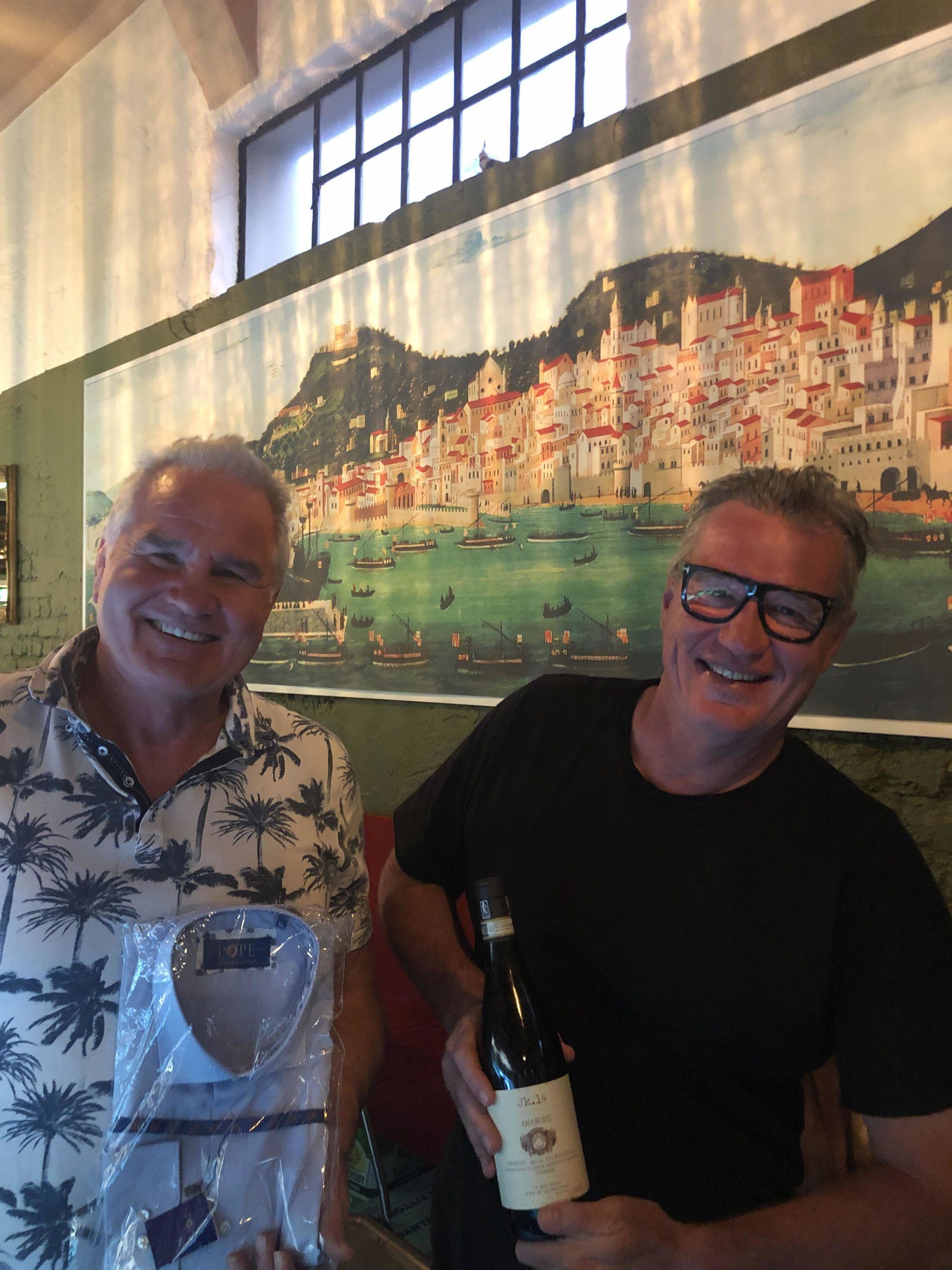 Two men smiling, one holding a shirt, the other a wine bottle, with Italian coastal painting backdrop.
