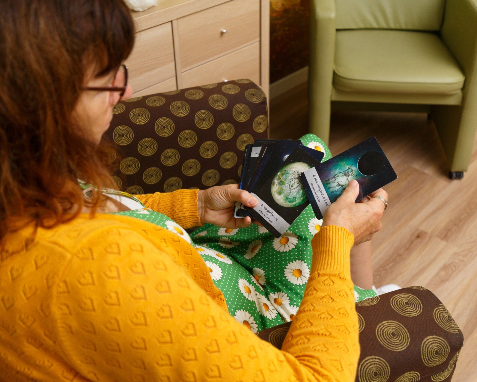Een vrouw in een gele trui en een bloemenjurk leest tarotkaarten op een stoel met patroon.