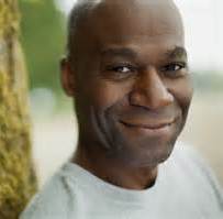 Smiling Black man wearing a gray shirt, leaning against a tree, outdoors.