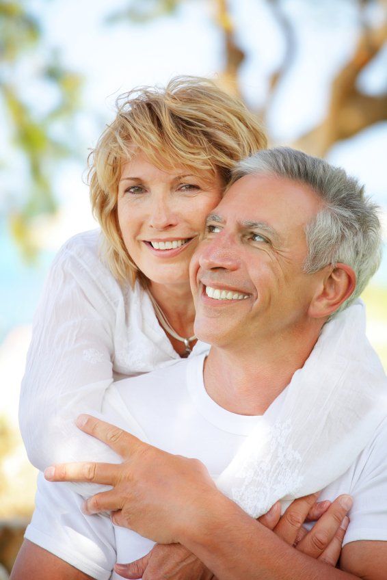 A smiling older couple embraces outdoors, with the woman's arms around the man's shoulders; both wear white.