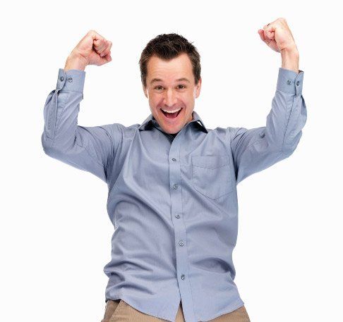 Man with short brown hair, light blue button-down shirt, tan pants, arms raised in a celebratory pose, smiling broadly.