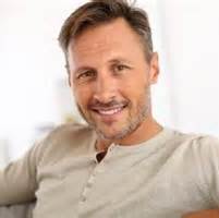 A smiling middle-aged man with brown hair and a light beard, wearing a beige Henley shirt, indoors.