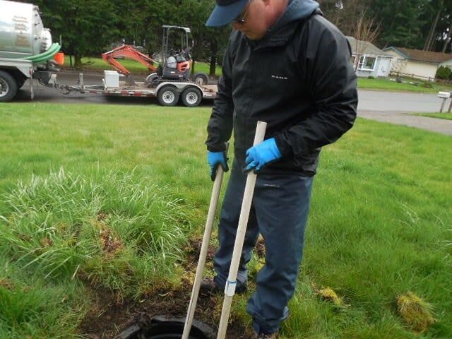 Man inspecting a septic tank, using sticks, in a grassy yard with service truck in background.
