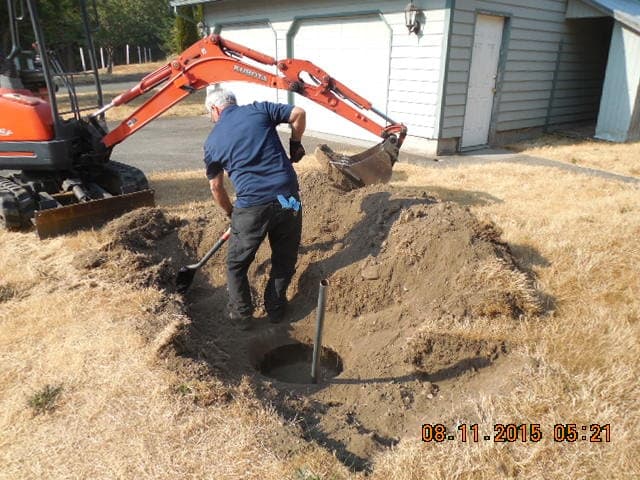 Man digging near a hole in the ground with an excavator nearby in front of a garage on a sunny day.