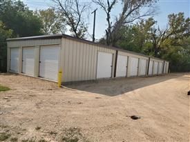 Storage units in a tan metal building with white doors, gravel driveway, and trees in the background.