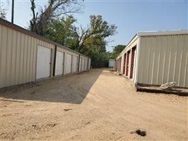Storage units in a row, beige metal buildings, dirt path, blue sky, and trees.
