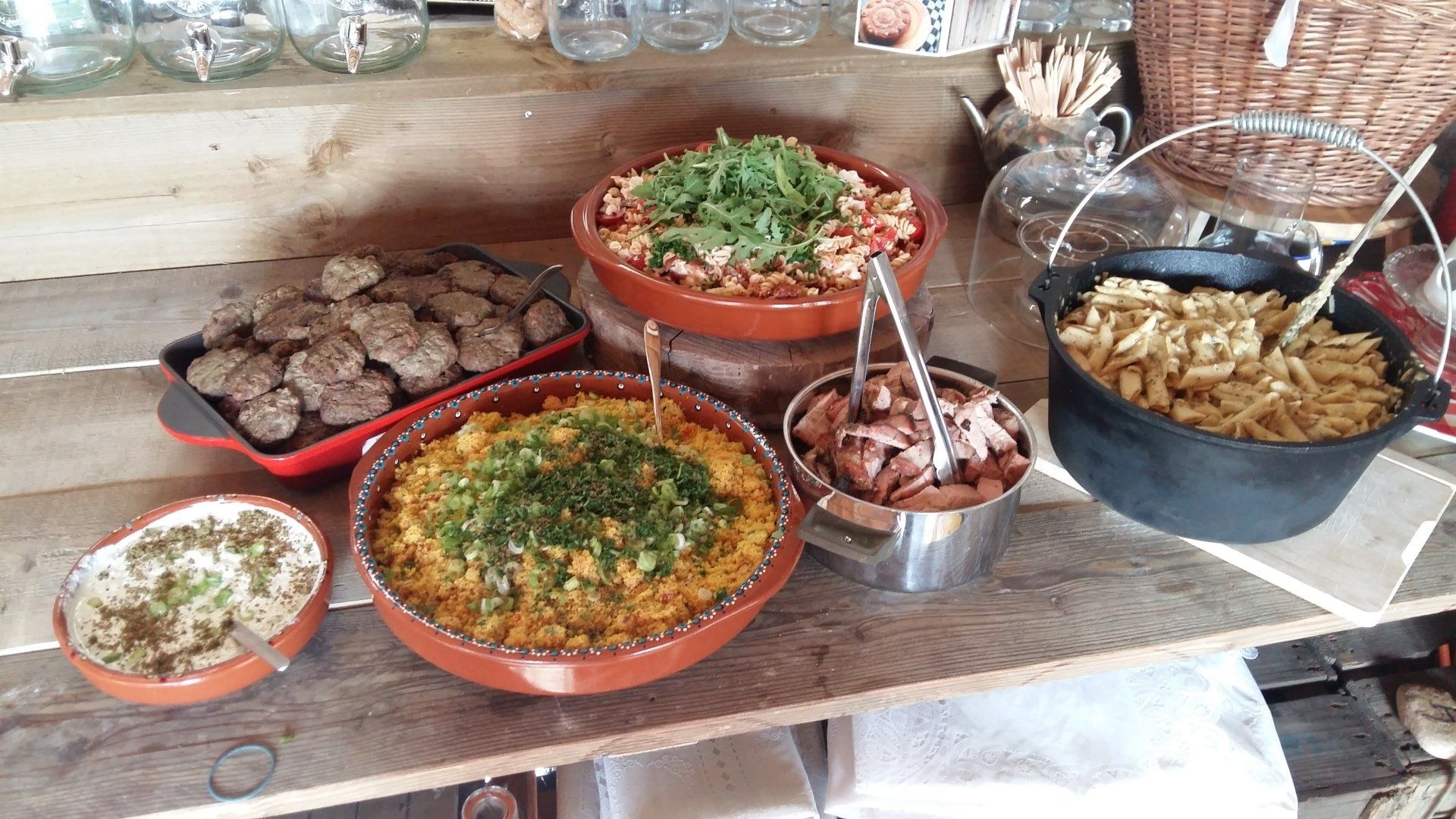 Buffet met diverse gerechten: gehaktballen, couscous, pasta, salades en bijgerechten op een houten tafel.