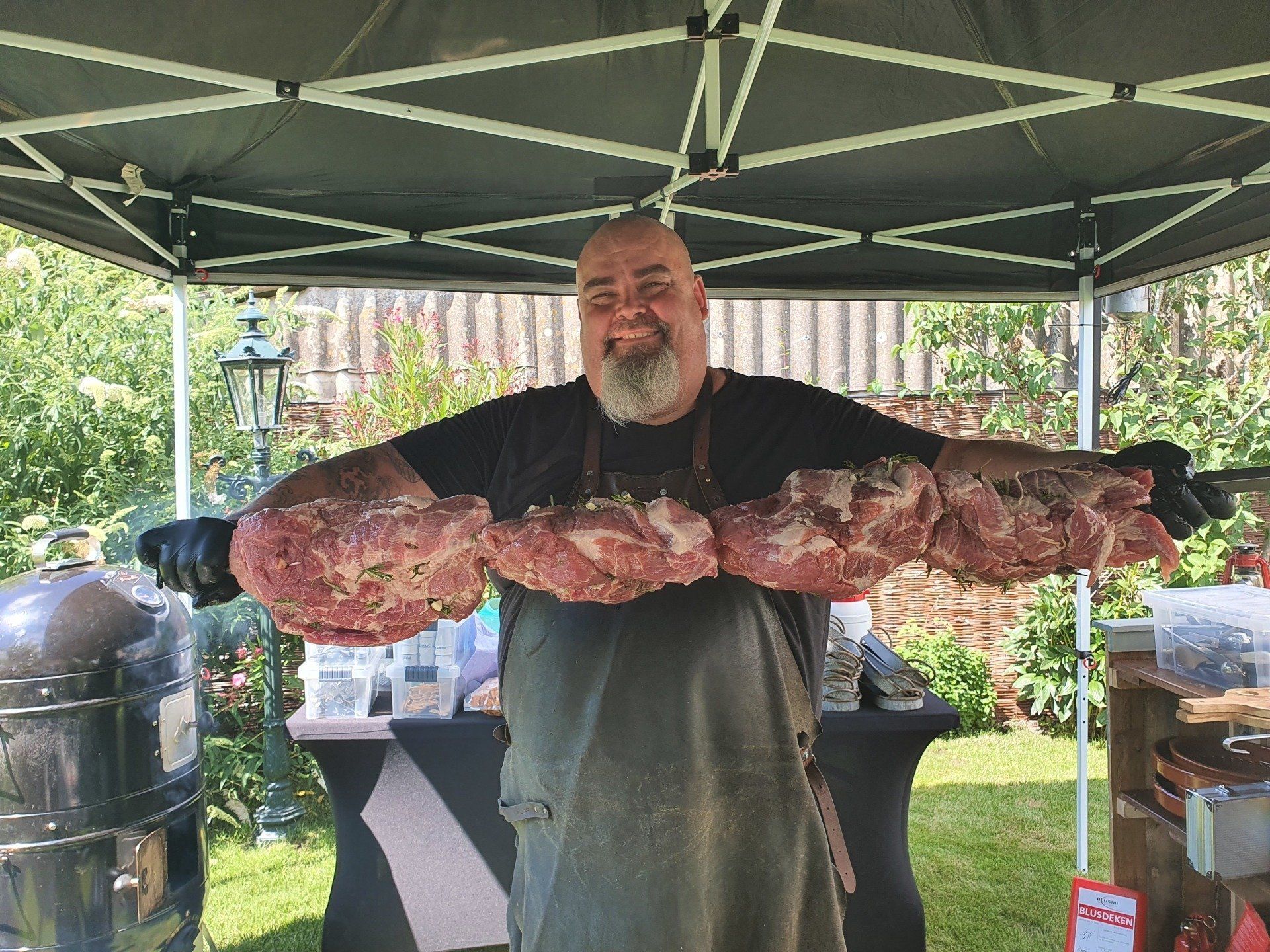 Chefkok Martin met grote stukken vlees staat lachend voor een rookoven en een tent, in een zonnige buitenomgeving.
