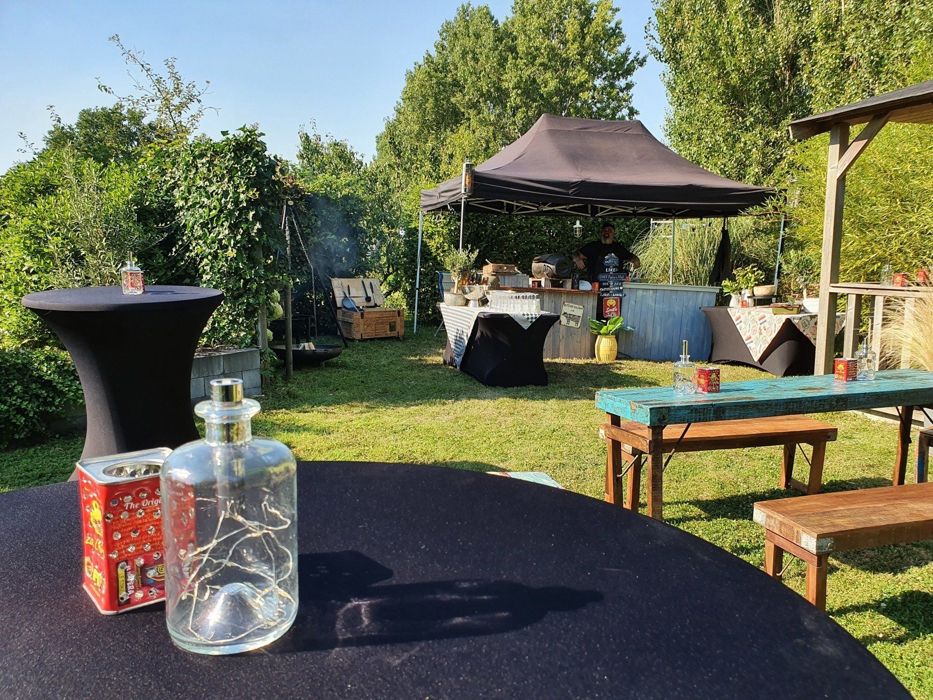 Feestscène in de buitenlucht: tafels met versieringen, een buffet onder een afdak, weelderig groen op de achtergrond.