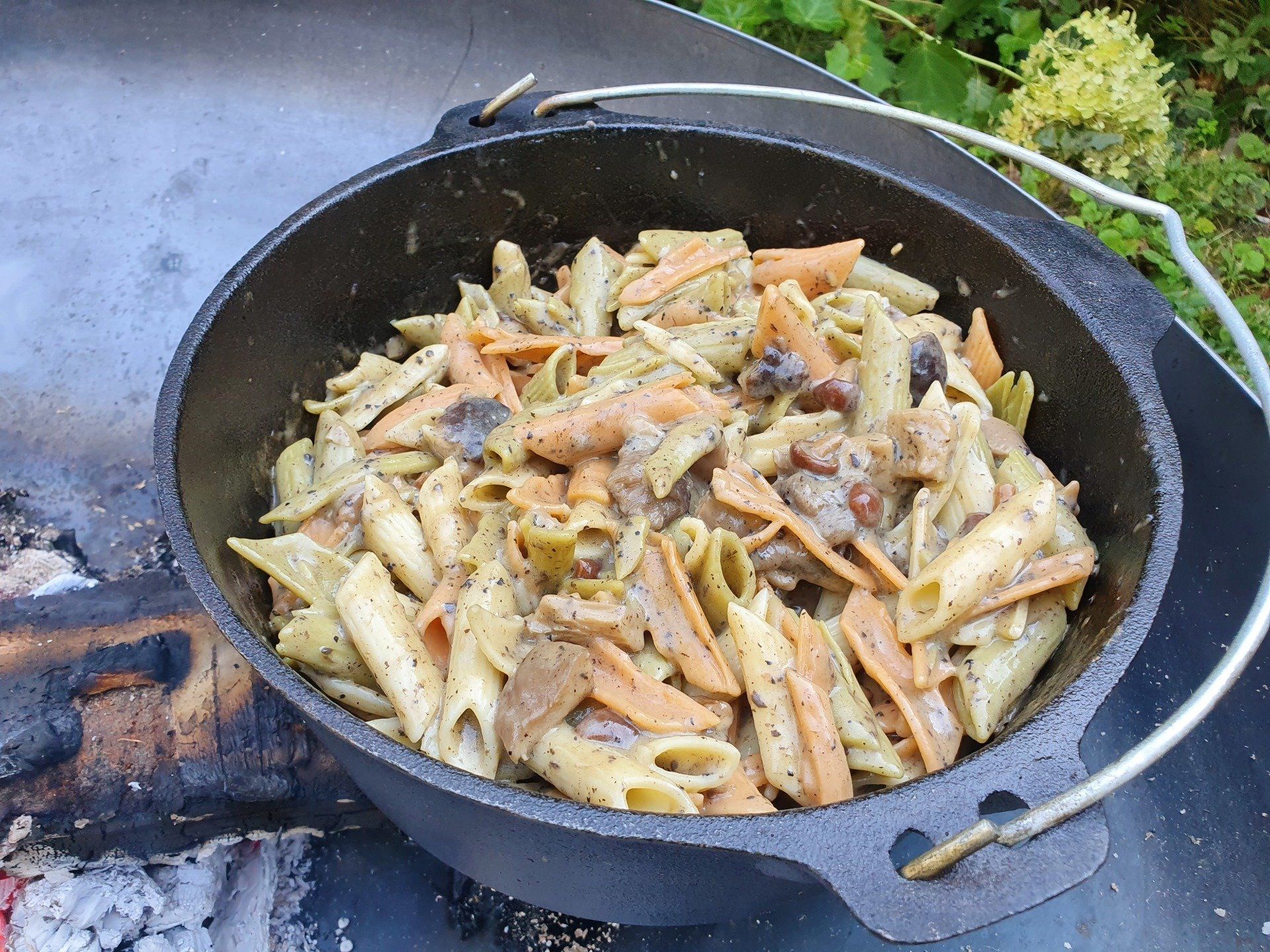 Pasta in een gietijzeren pot boven een kampvuur, met champignons en kleurrijke pastavormen.