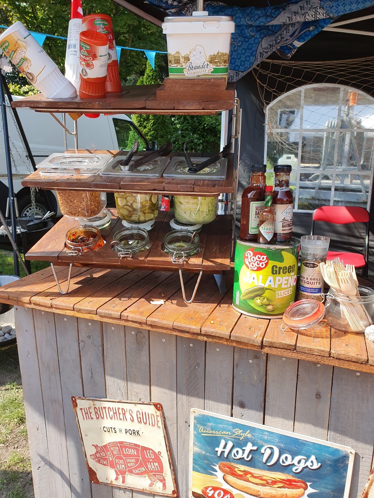 Hotdogkraam met sauzen en toppings op houten planken.