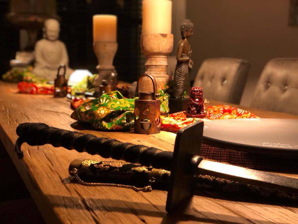 Een tafel gedekt met zwaarden en kaarsen; een Boeddhabeeld, decoratieve objecten en borden zijn zichtbaar.