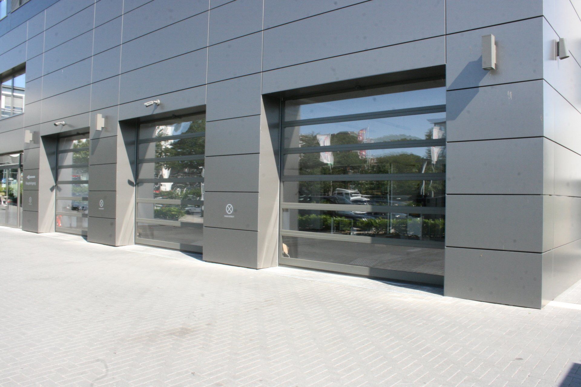 Moderne Gebäudefassade mit drei Garagentoren aus Glas. Graue Paneele und Pflasterung. Sonnenlicht.