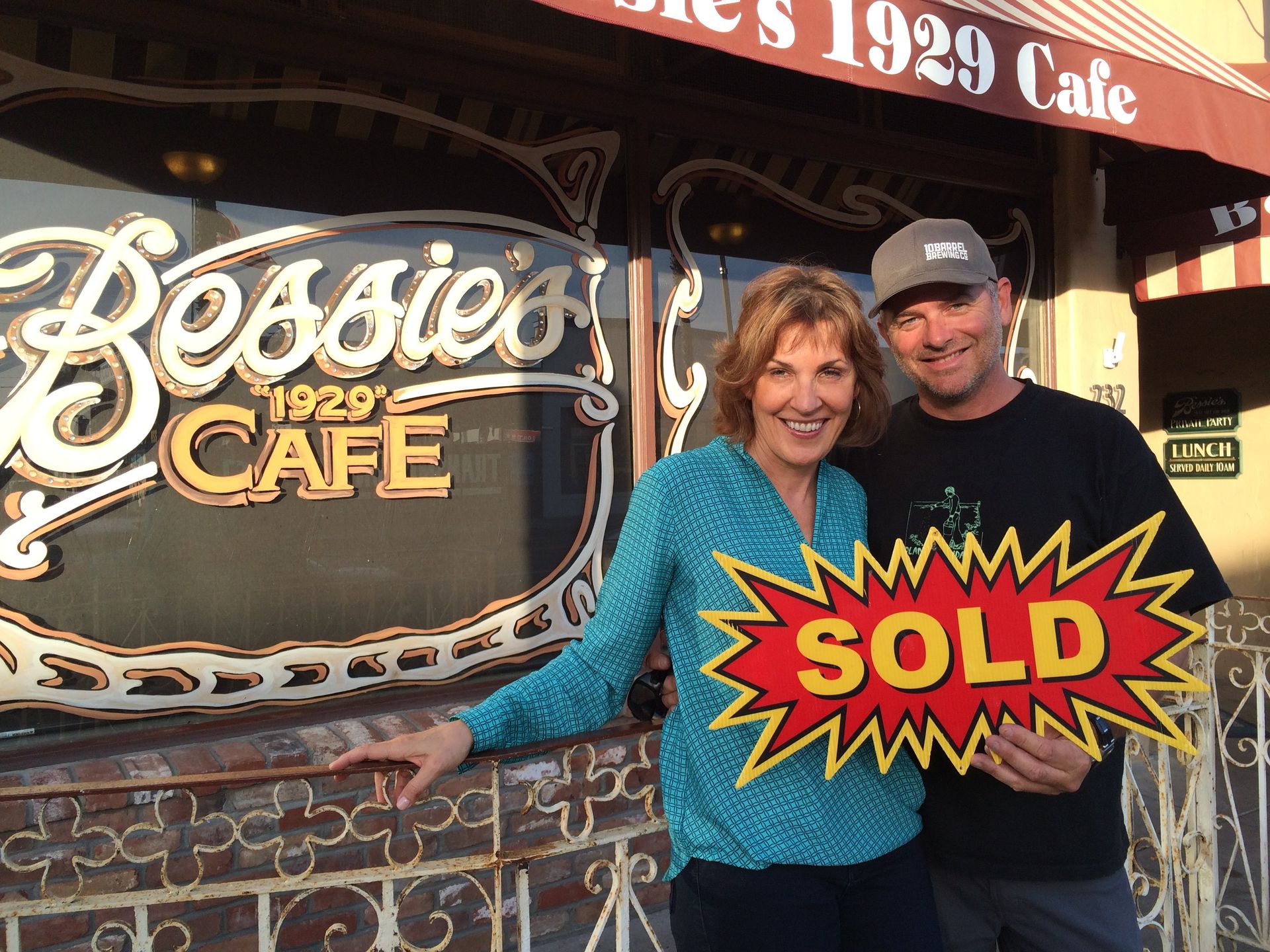 Couple posing outside Bevo’s 1929 Cafe, holding a bright red and yellow SOLD sign