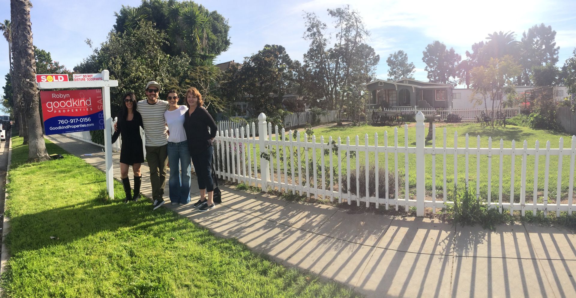 People standing by a white picket fence near a park sign on a sunny day