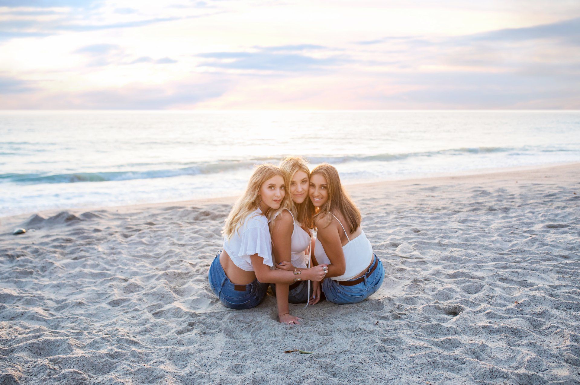 Three women are sitting on the beach hugging each other.