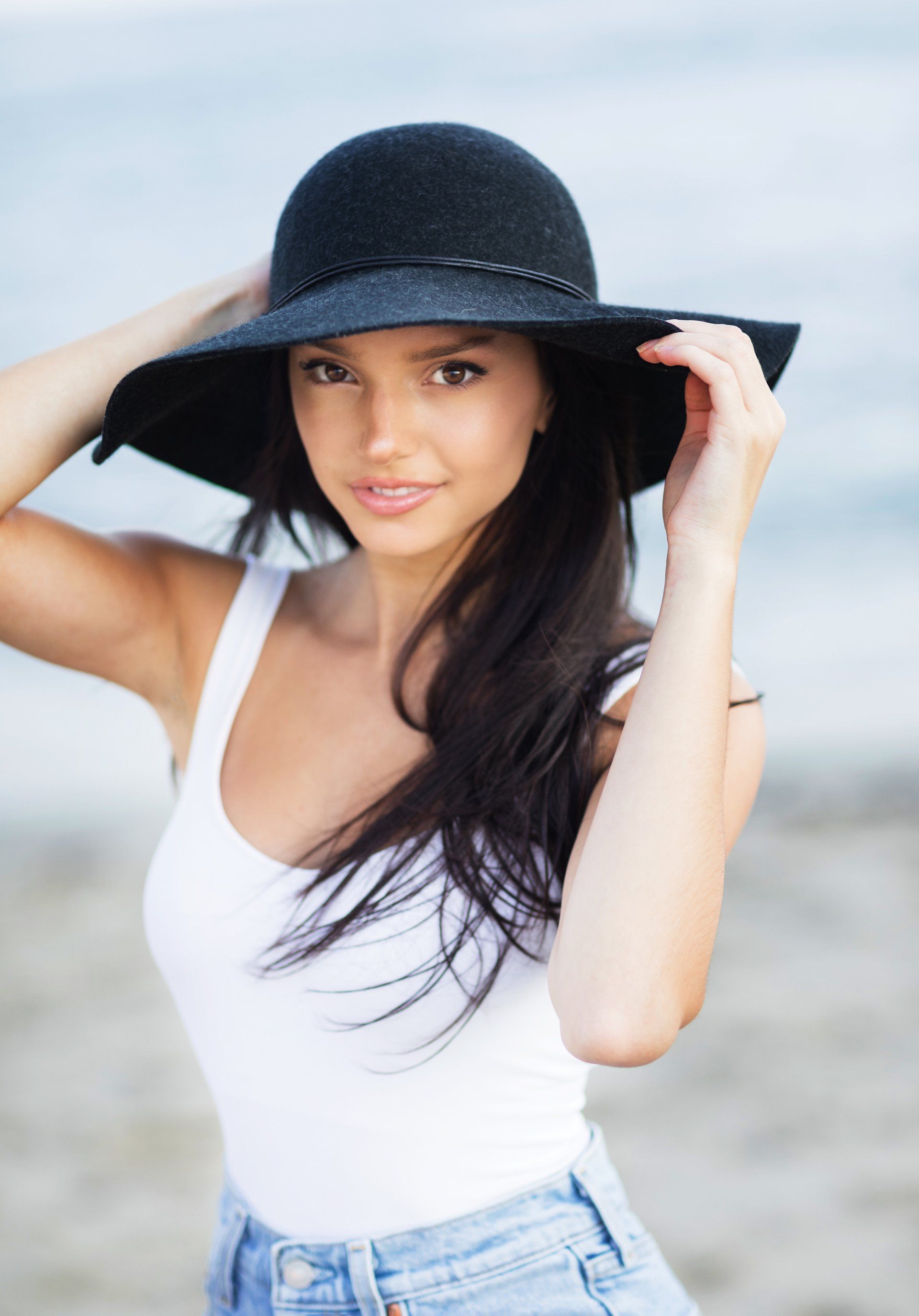 A woman wearing a black hat and a white tank top is standing on the beach.