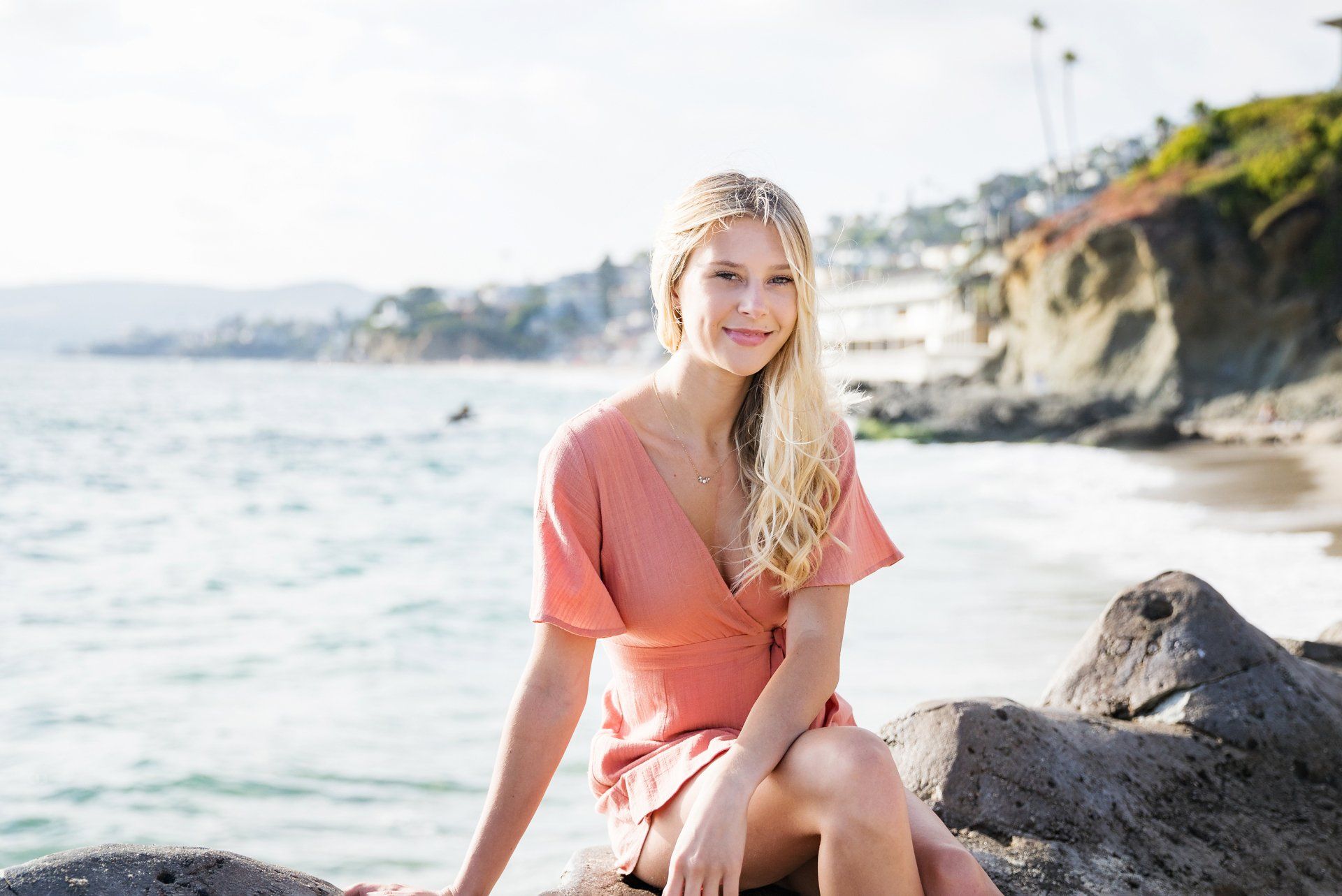 A woman in a pink dress is sitting on a rock near the ocean.