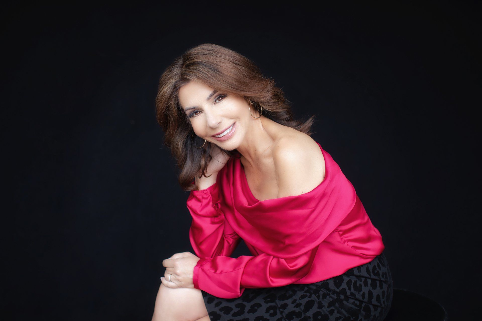 A woman in a red dress is sitting on a stool with her legs crossed and smiling.