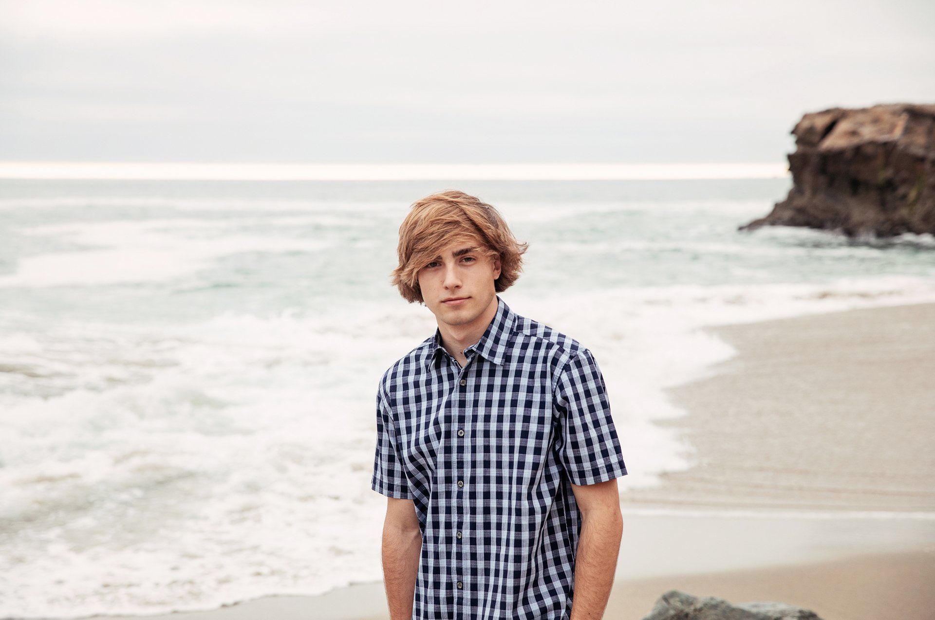 A young man in a plaid shirt is standing on a beach.