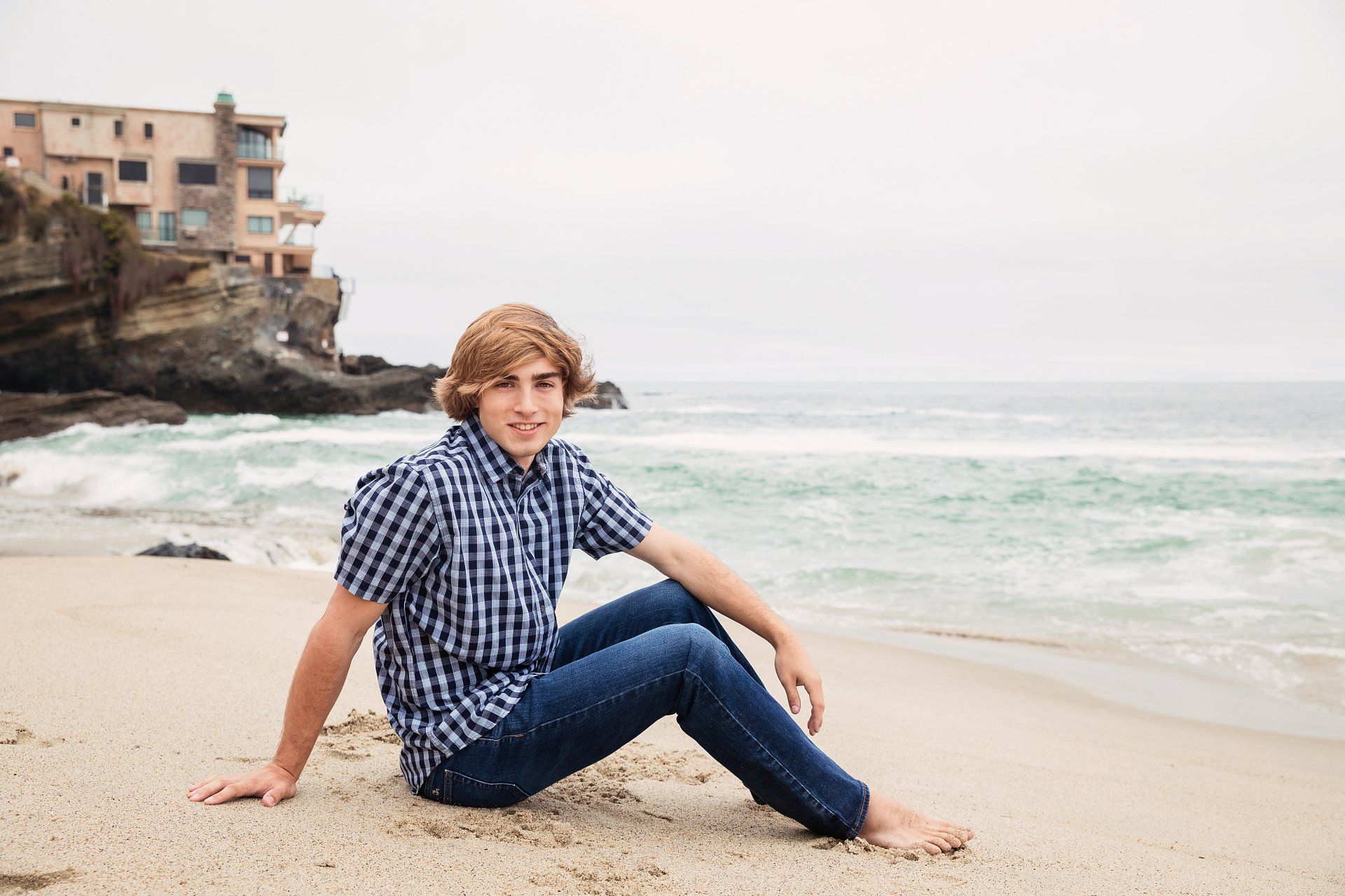 A young man is sitting on the beach near the ocean.
