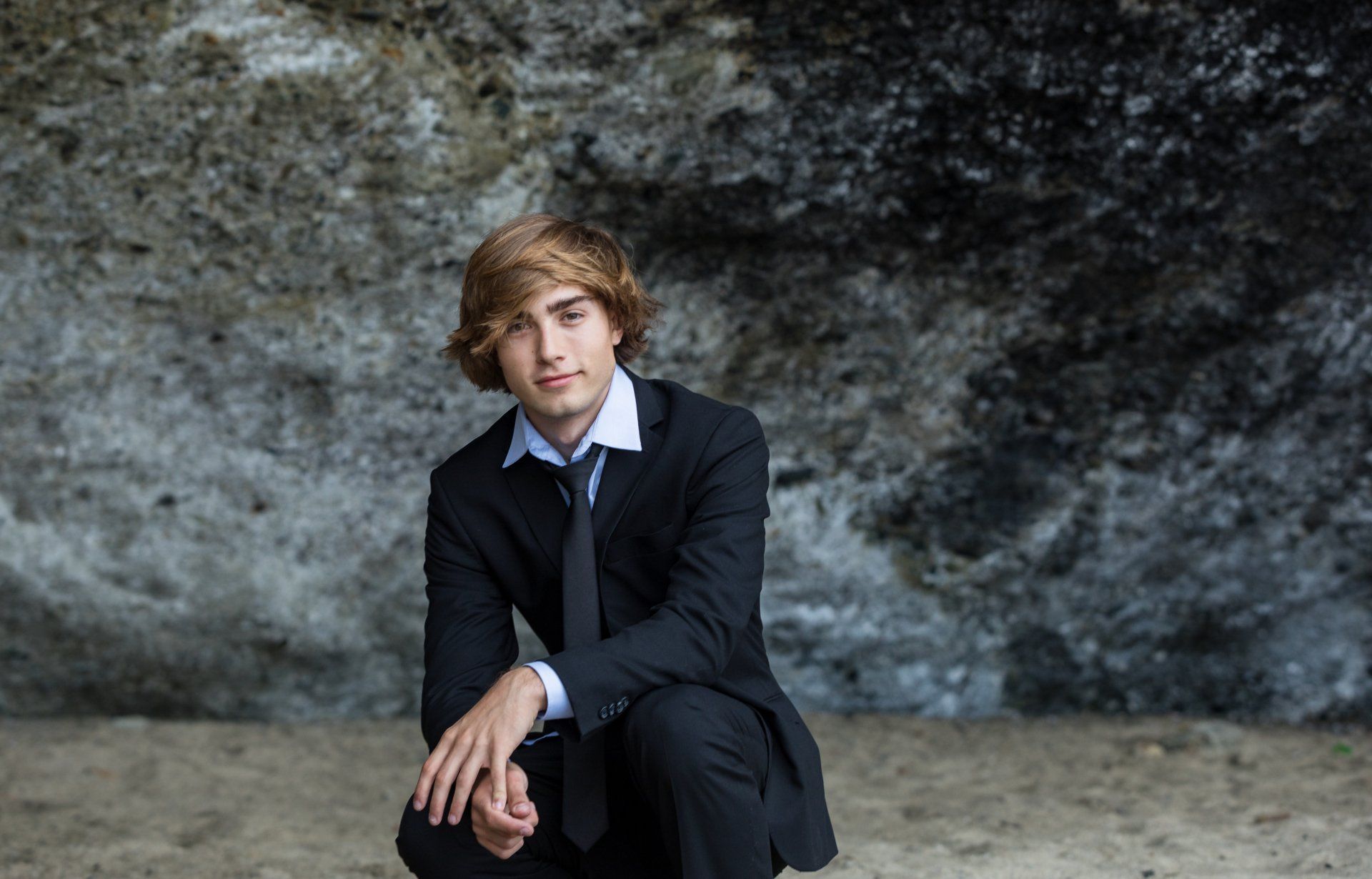 A young man in a suit and tie is kneeling down in front of a rock wall.