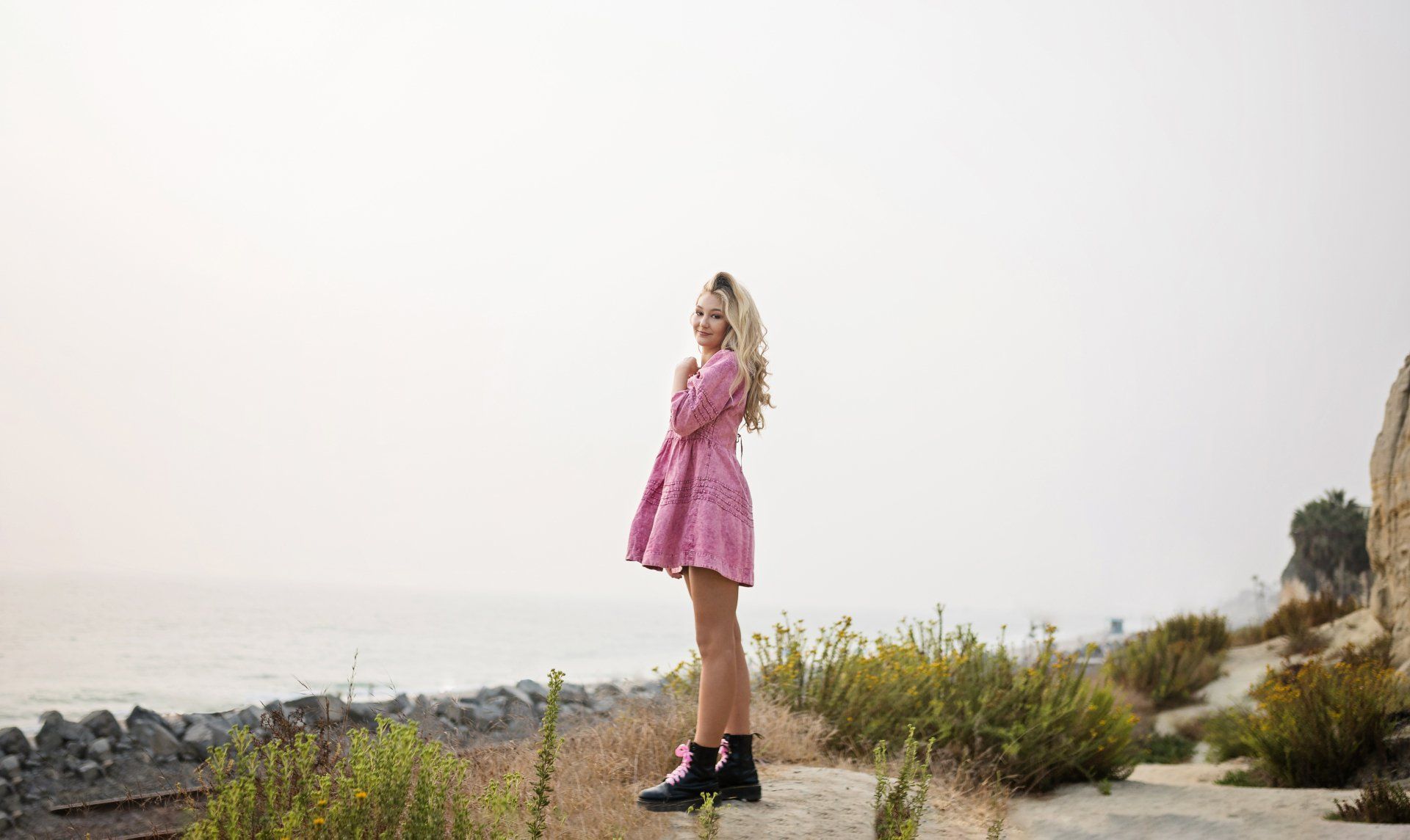 A woman in a pink dress is standing on top of a hill overlooking the ocean.