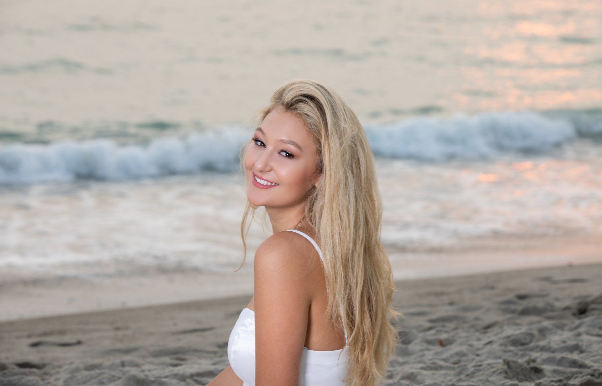 A woman in a white top is sitting on the beach looking at the camera.