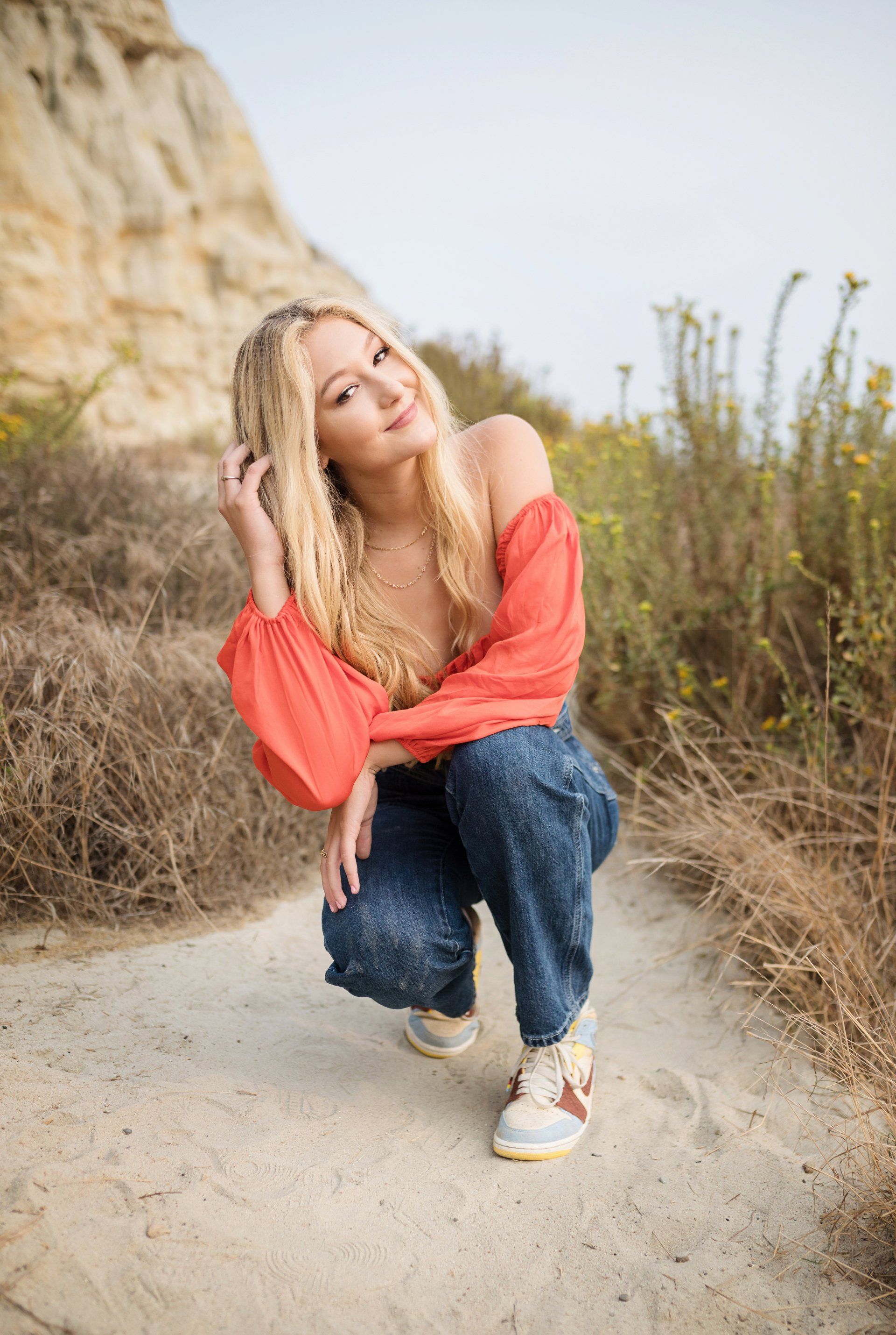 A woman in a red top and jeans is squatting down on a dirt path.