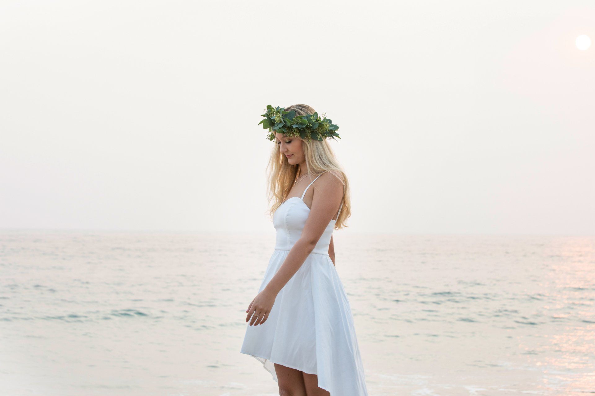 A woman in a white dress with a flower crown on her head is standing on the beach.