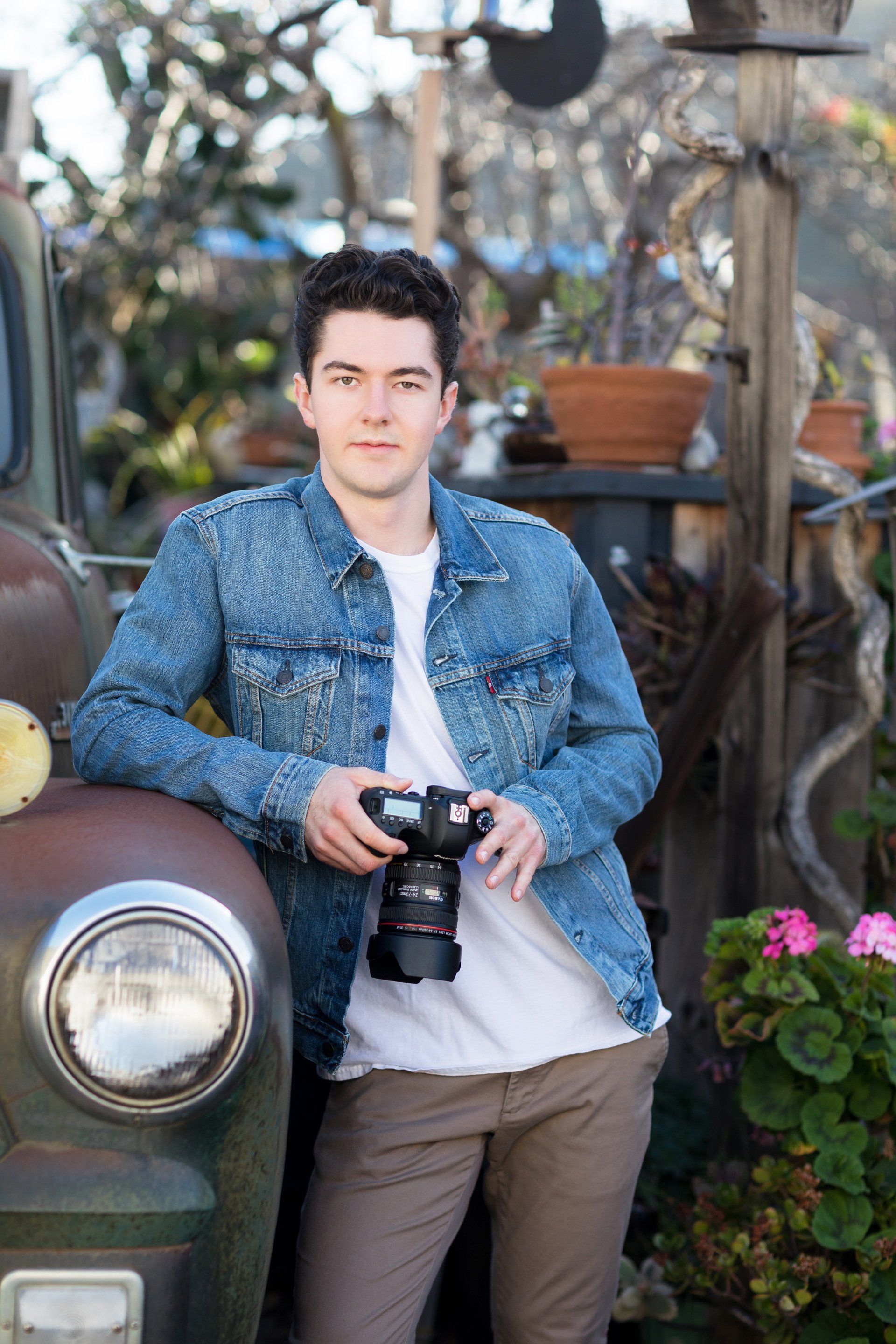 A young man in a denim jacket is standing next to an old truck holding a camera.