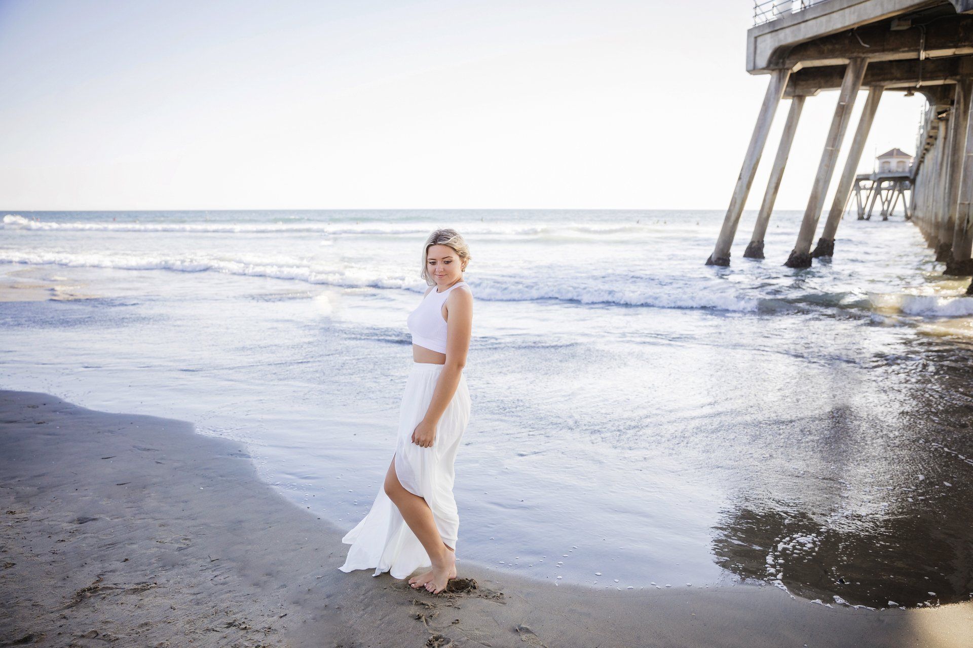 A woman in a white dress is standing on the beach near a pier.