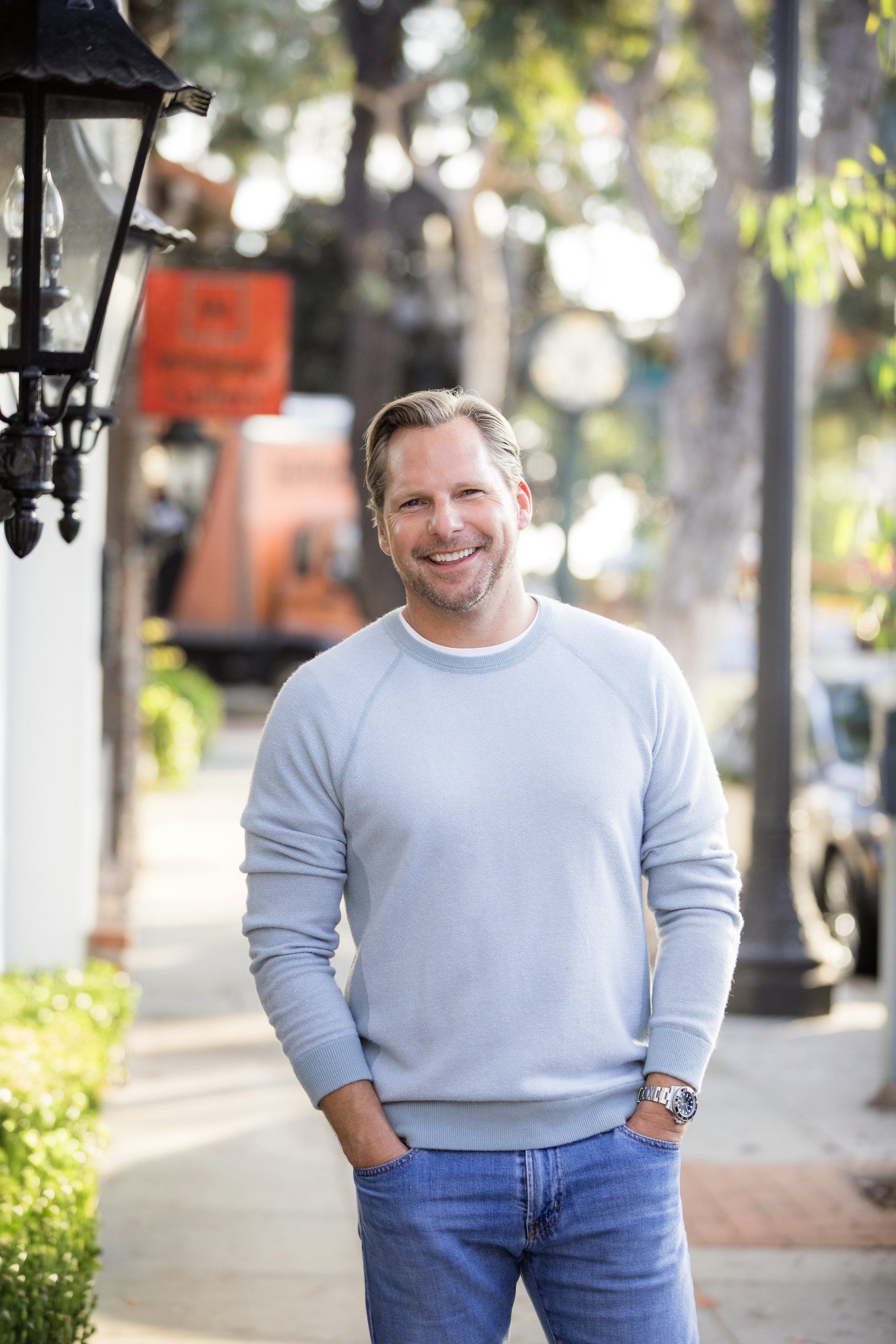 A man in a grey sweater and blue jeans is standing on a sidewalk with his hands in his pockets.