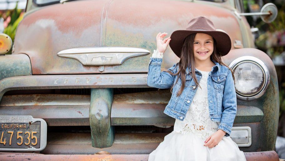 A little girl wearing a hat and a denim jacket is sitting on the front of an old truck.