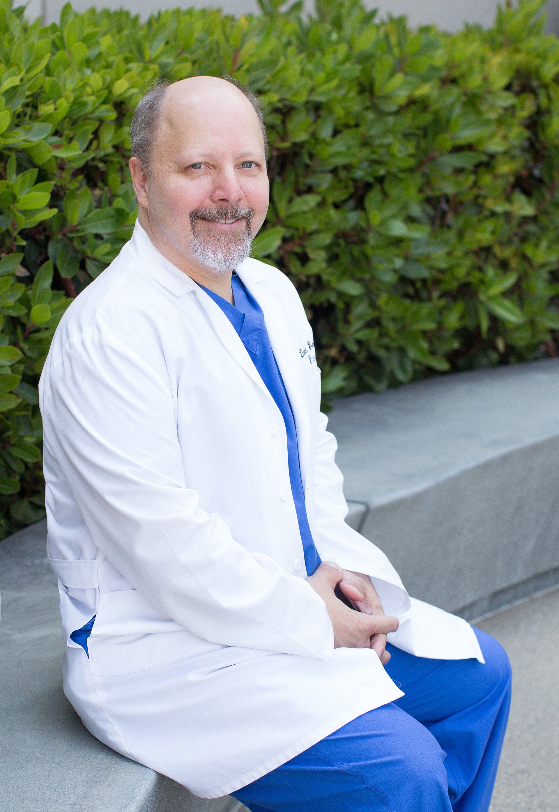A man in a white lab coat and blue scrubs is sitting on a bench.