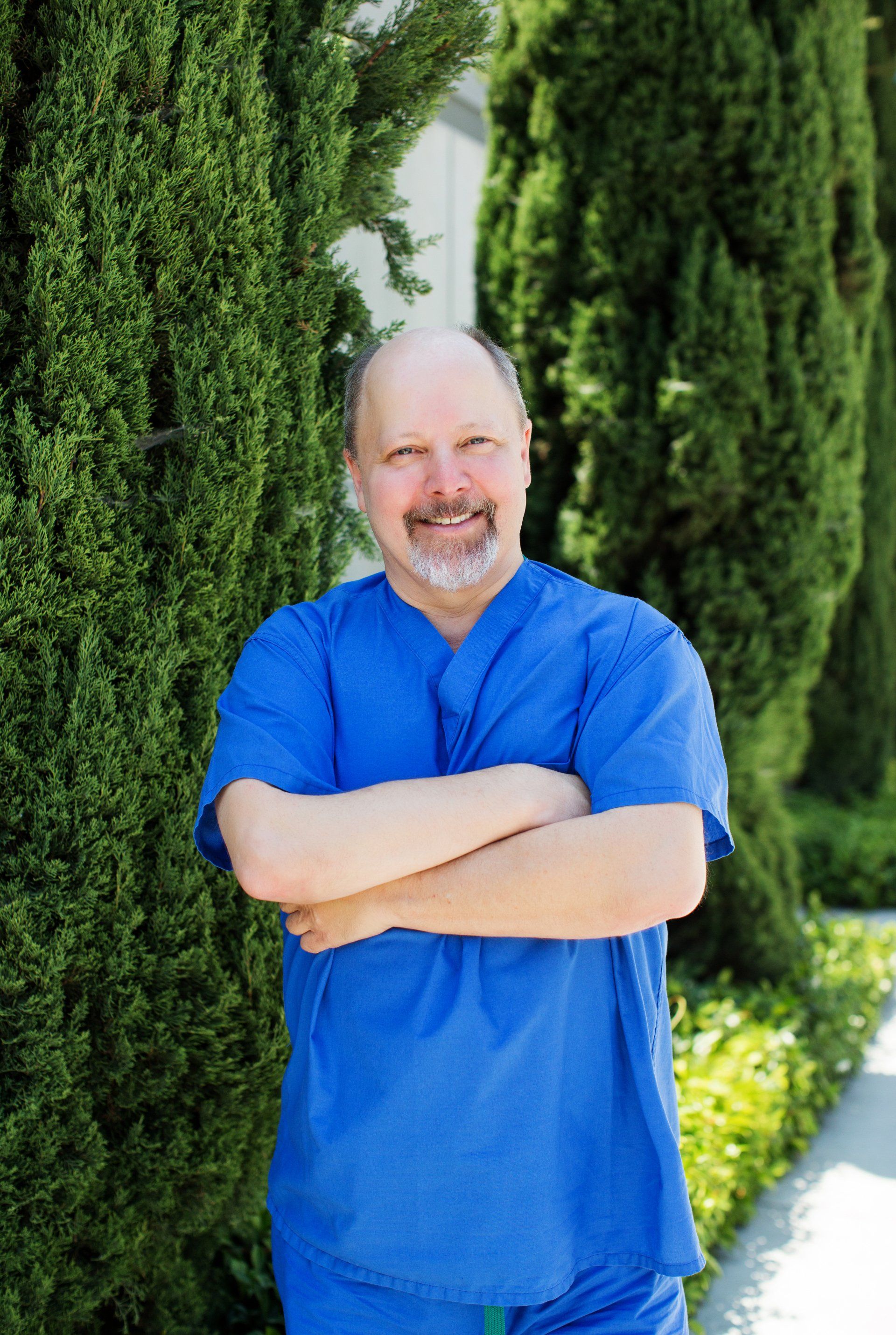 A man in a blue scrub is standing with his arms crossed in front of trees.