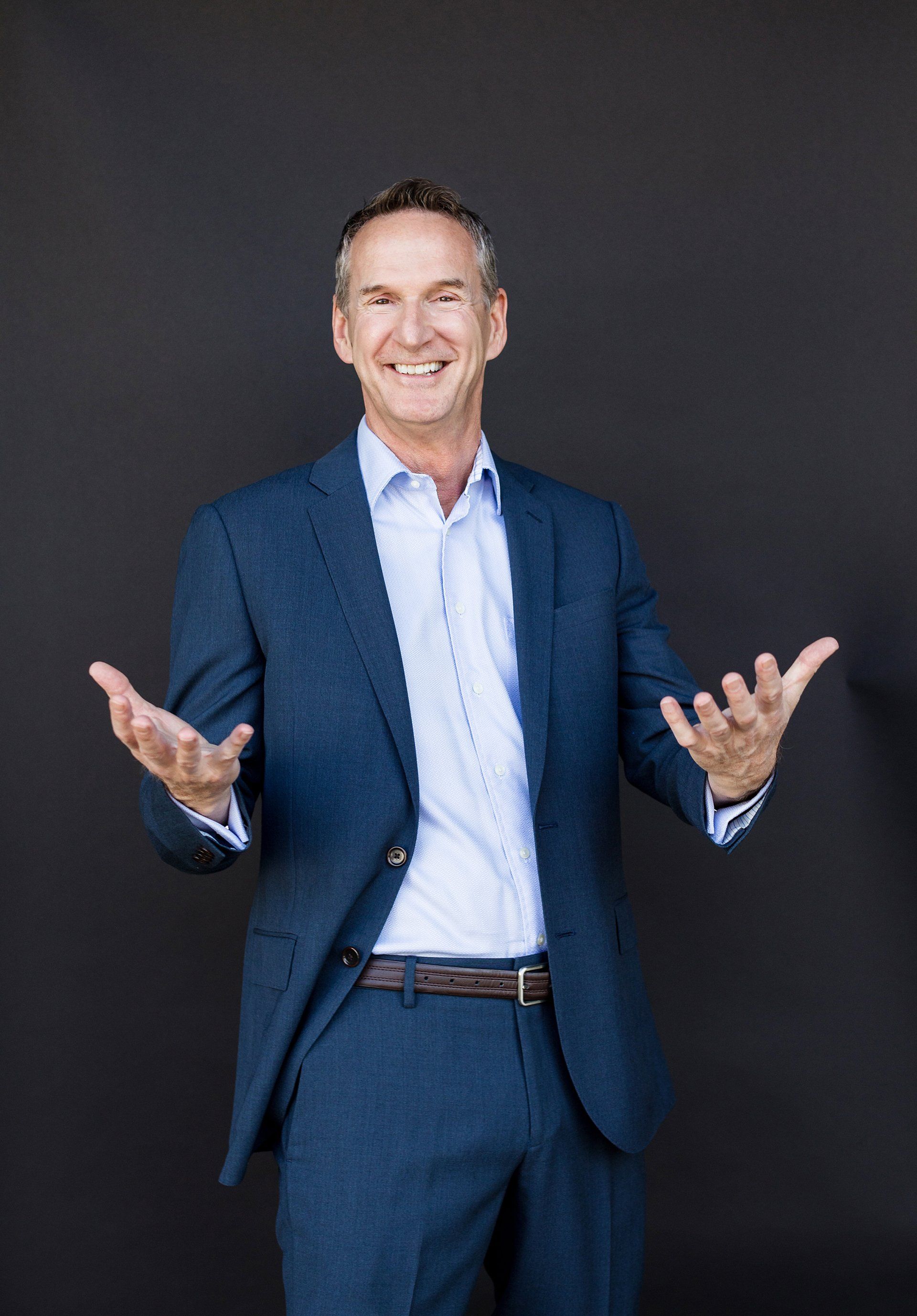 A man in a blue suit and white shirt is standing with his arms outstretched.