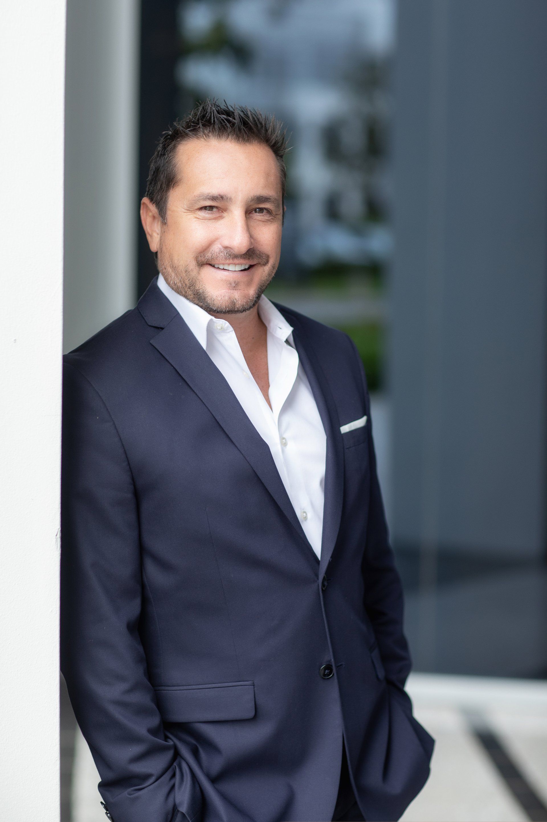 A man in a suit is leaning against a wall with his hands in his pockets.