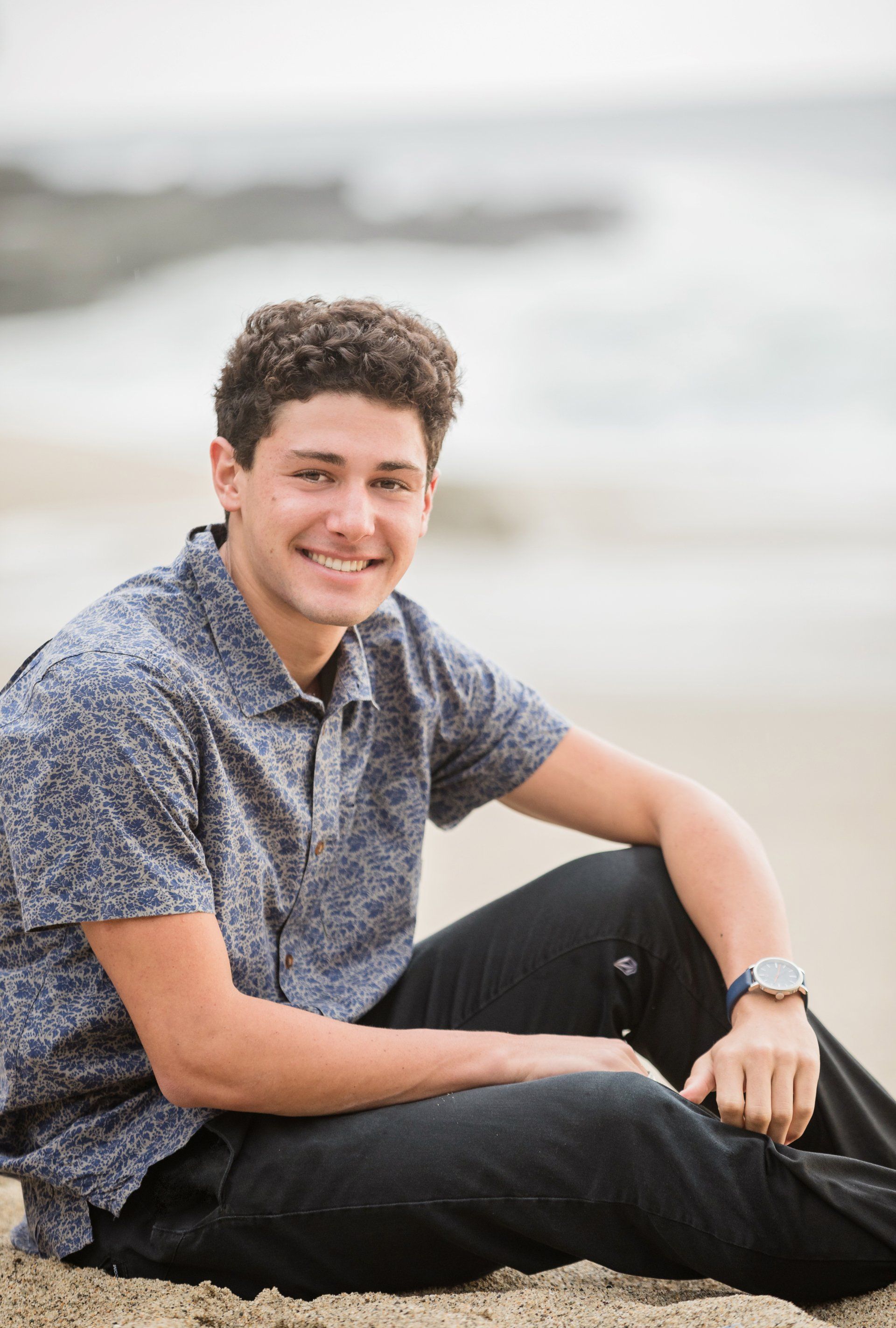 A young man is sitting on the beach wearing a watch and smiling.